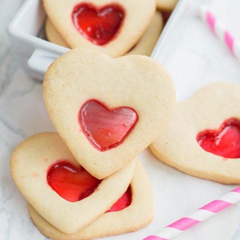Close up view of heart shaped stained glass cookies stacked on white marble counter in front of small white serving dish. Pink and white striped paper straws laying nearby.