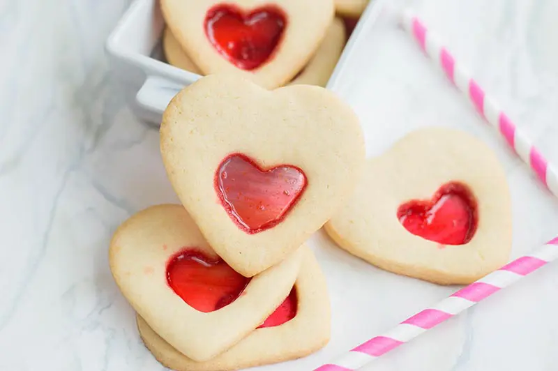 Close up view of heart shaped stained glass cookies stacked on white marble counter in front of small white serving dish. Pink and white striped paper straws laying nearby.