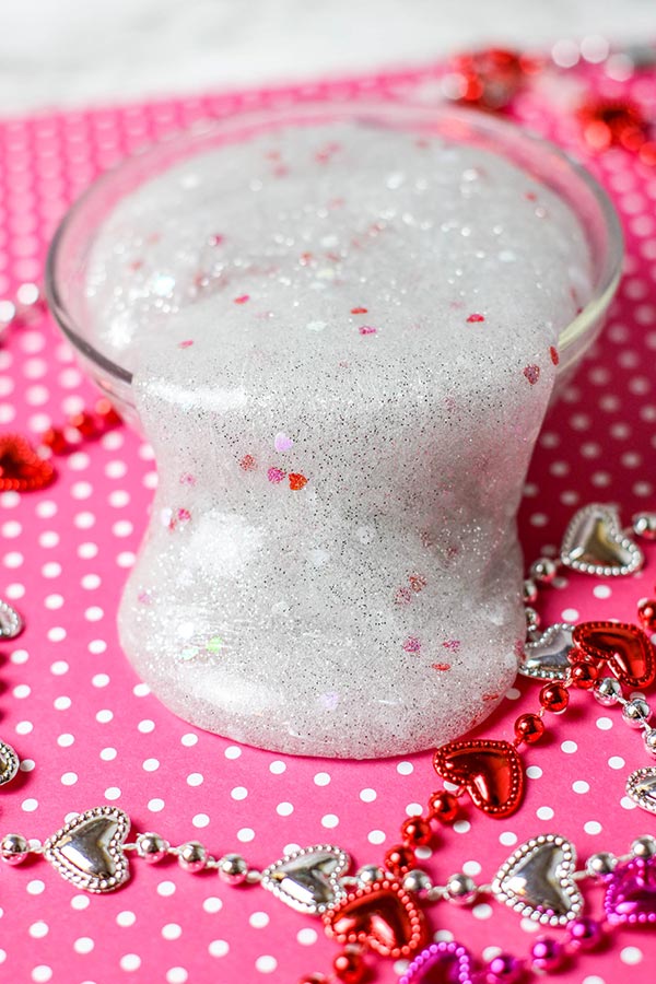 Shiny slime with silver glitter and little red and pink hearts spilling over side of small round container on top of pink paper with white polka dots. Shiny heart necklaces around it.