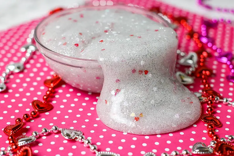Slime with glitter and heart confetti in small round bowl spilling over the side onto pink paper with white dots.