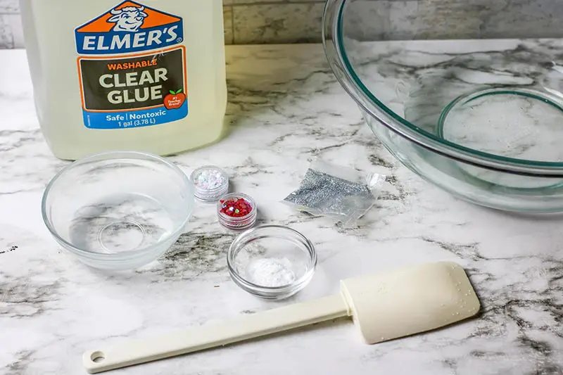 Jug of clear glue, clear bowls, glitter, and heart confetti on counter.