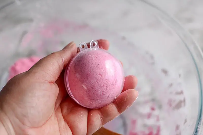 Hand holding closed round bath bomb mold filled with pink bath bomb ingredients.