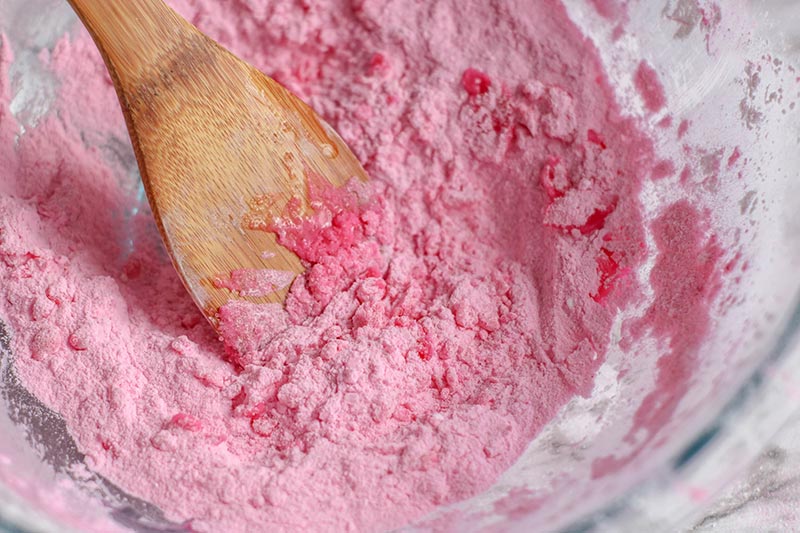 Pink bath bomb ingredients being mixed together in glass bowl with wooden spoon.