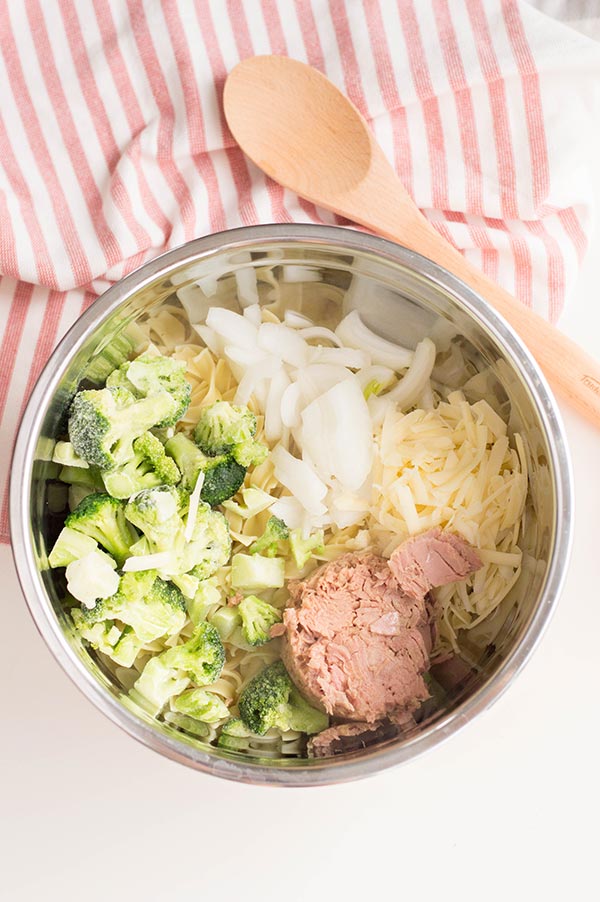 Broccoli, onion, shredded cheese, canned tuna fish in silver bowl next to wood spoon on red and white kitchen cloth.