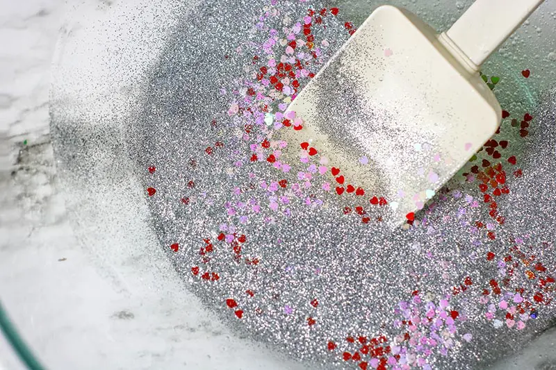 White spatula mixing silver glitter, red foil heart confetti, and pink foil heart confetti into glue mixture.