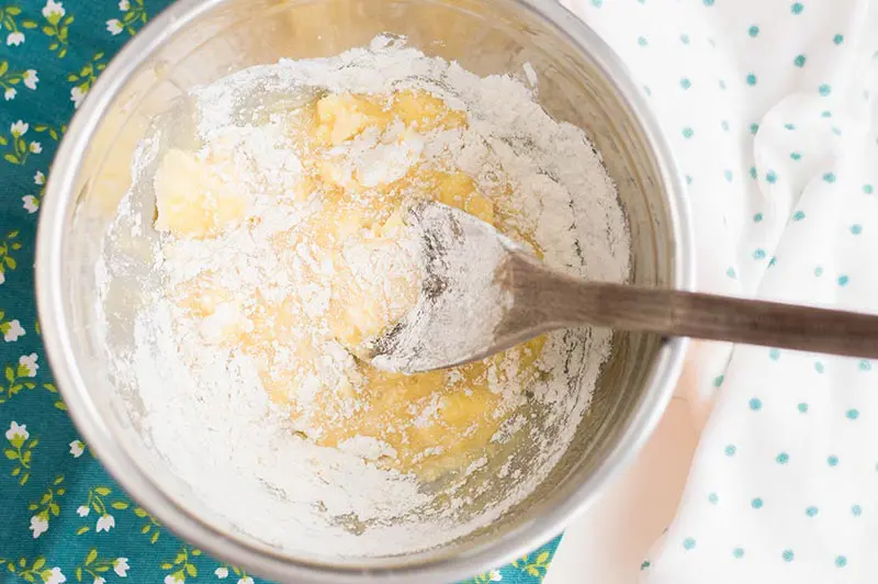 Mixing sugar cookie dough ingredients in silver mixing bowl with wooden spoon.