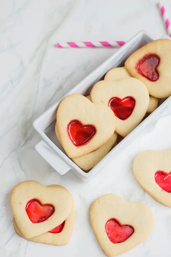 Heart shaped sugar cookies with melted candy centers arranged on white marble counter top and in small white rectangular serving dish.