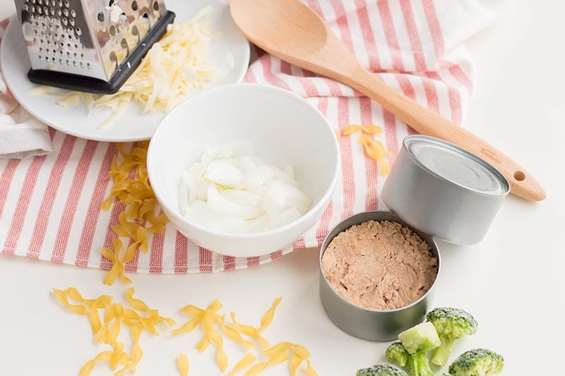 Open can of tuna, broccoli, and egg noodles on white counter. Chopped onion in small white bowl. Cheese grater and shredded cheese and wooden spoon on red and white striped kitchen towel.