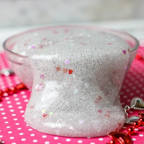 Close up of silver glitter slime with heart confetti flowing over side of small clear bowl.