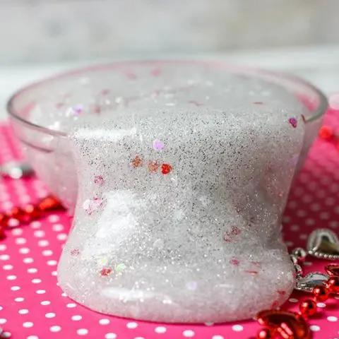 Close up of silver glitter slime with heart confetti flowing over side of small clear bowl.