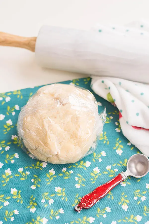 Sugar cookie dough in a ball and wrapped in plastic. Ball of dough is siting on blue kitchen towel with little flowers and next to a marble rolling pin.