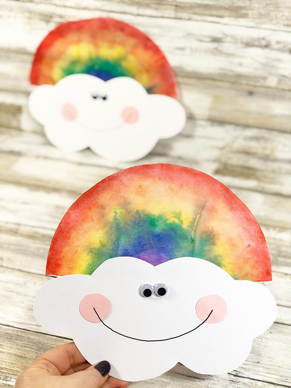 Woman's hand holding finished rainbow colored coffee filter with cute smiling cloud face. Another completed project is laying down in the background.