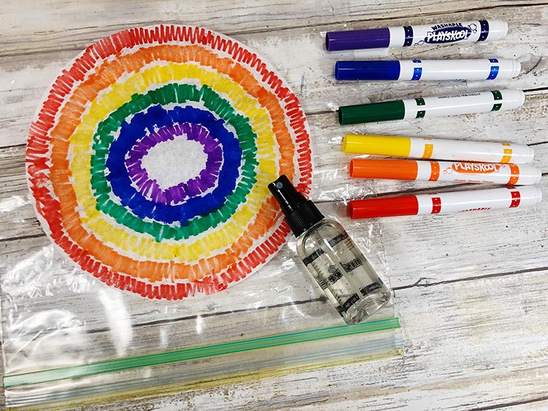 Flattened coffee filter drawn on with markers in rainbow color order. Markers laying next to filter and small spray bottle with water.