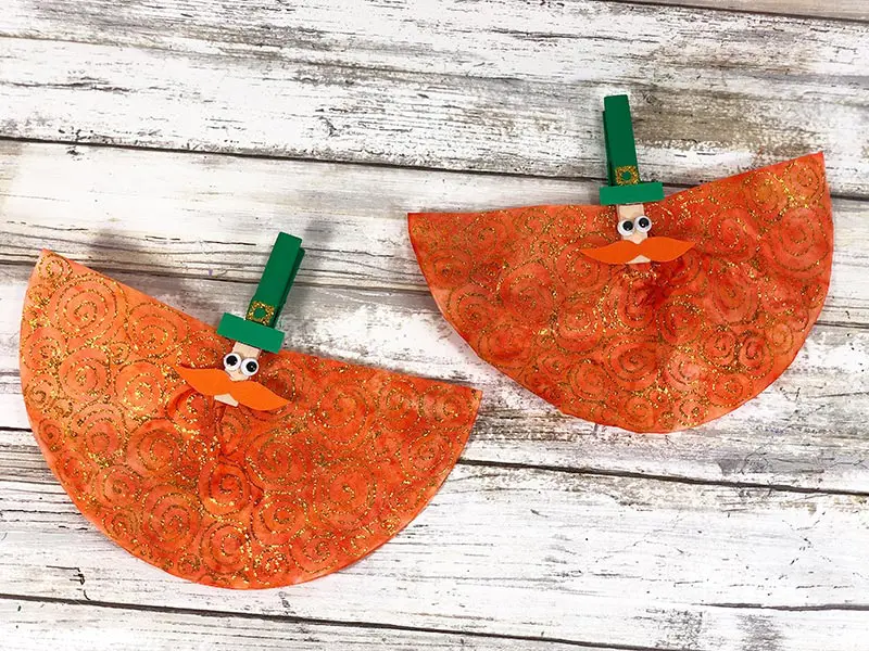 Two leprechauns made with clothespins and coffee filters laying on white wood background.
