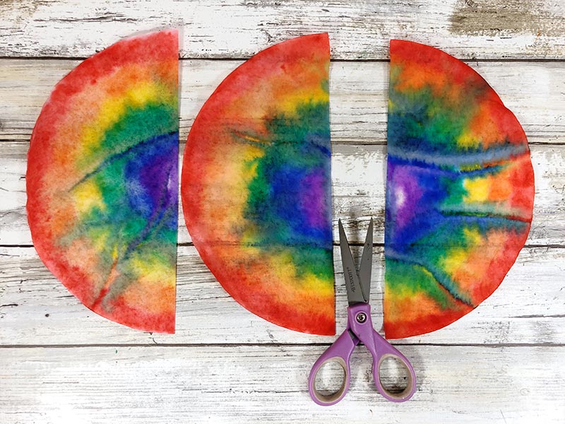 Rainbow colored coffee filters cut in half laying on white wood background with pair of purple handled scissors.