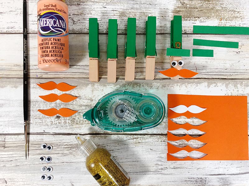 Painted clothespins lined up and orange paper mustaches cut out. Supplies to glue them together.