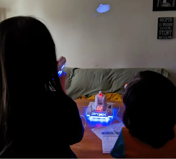 Two kids aiming ProjeX blasters at UFO target on white wall above couch.