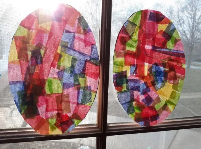 Two finished tissue paper Easter egg suncatchers hanging in sunny window.
