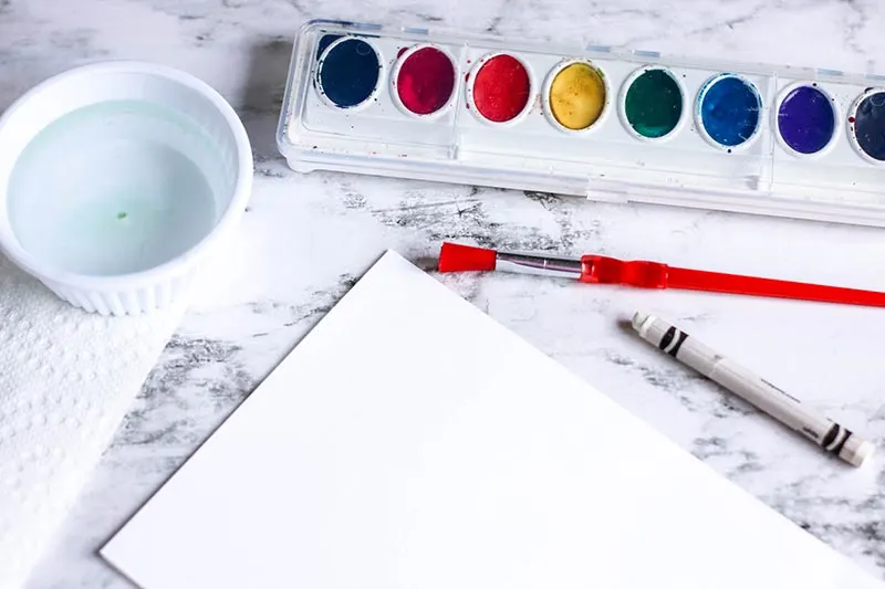 Watercolor paint set, paint brush, small cup of water, white crayon, and white paper folded in half on counter.