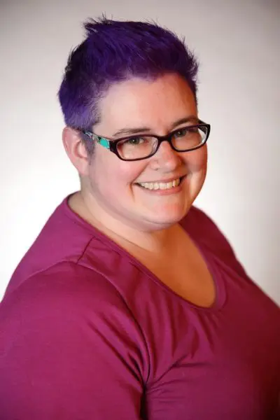 Headshot photo of white woman with short purple hair, glasses, smiling, wearing a dark pink top.