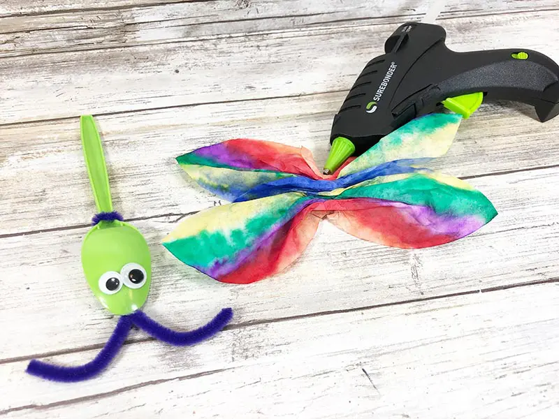 Green plastic spoon with googly eyes and purple chenille stem antenna laying on white wood background next to rainbow colored coffee filter wings and glue gun.