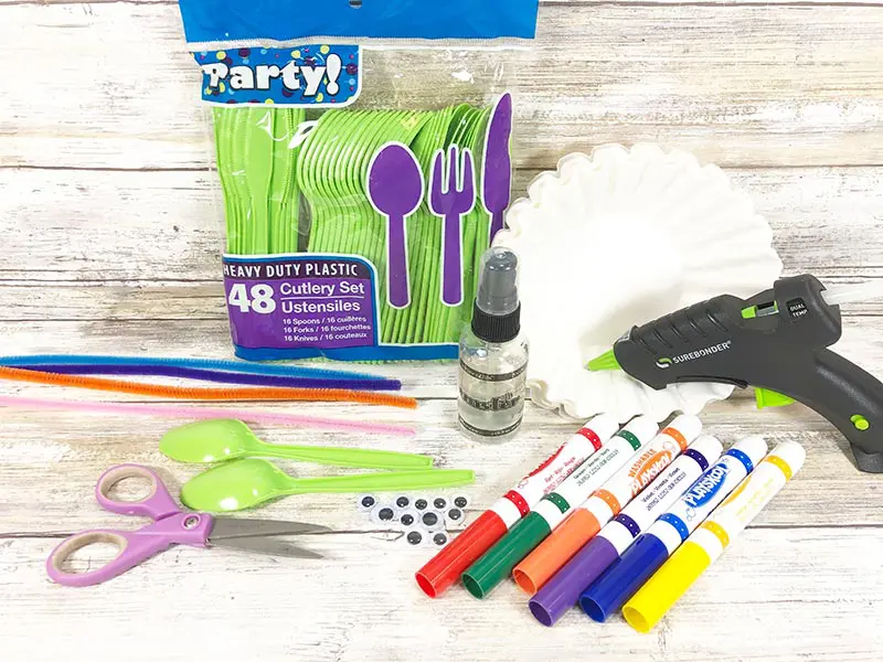 Pack of green plastic spoons, coffee filters, pipe cleaners, googly eyes, markers, scissors, and hot glue gun on white wood background.