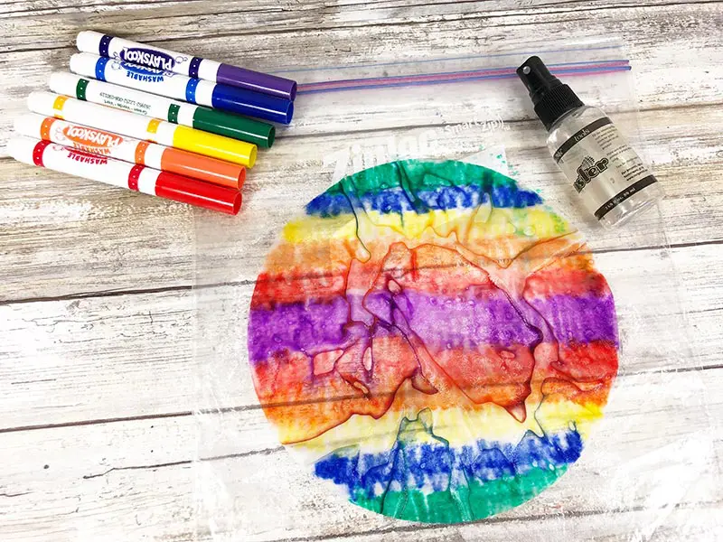 Wet colored coffee filter with colors blending, washable markers, and small water spray bottle laying on white wood background.