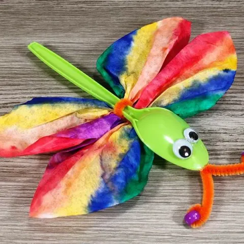 Close view of completed dragonfly craft made with coffee filter and plastic spoon on gray wood background.