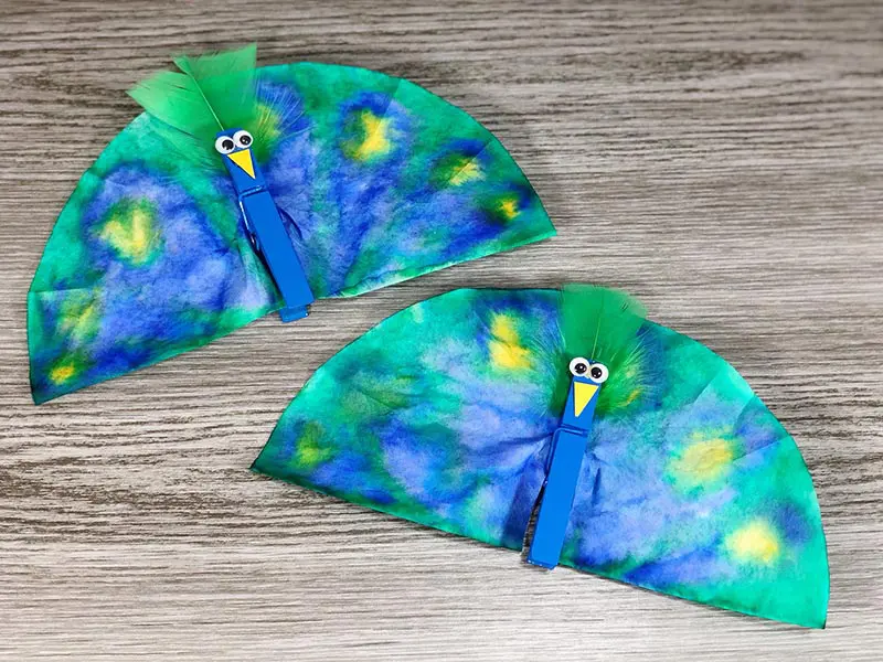 Two peacock craft projects laying next to each other on gray wood background.