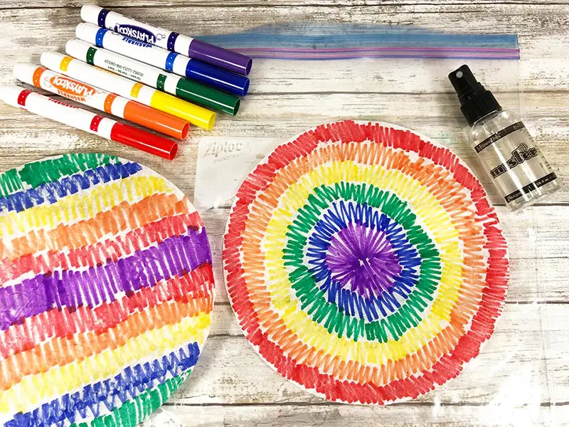 Two rainbow colored coffee filters and markers laying on white wood background.