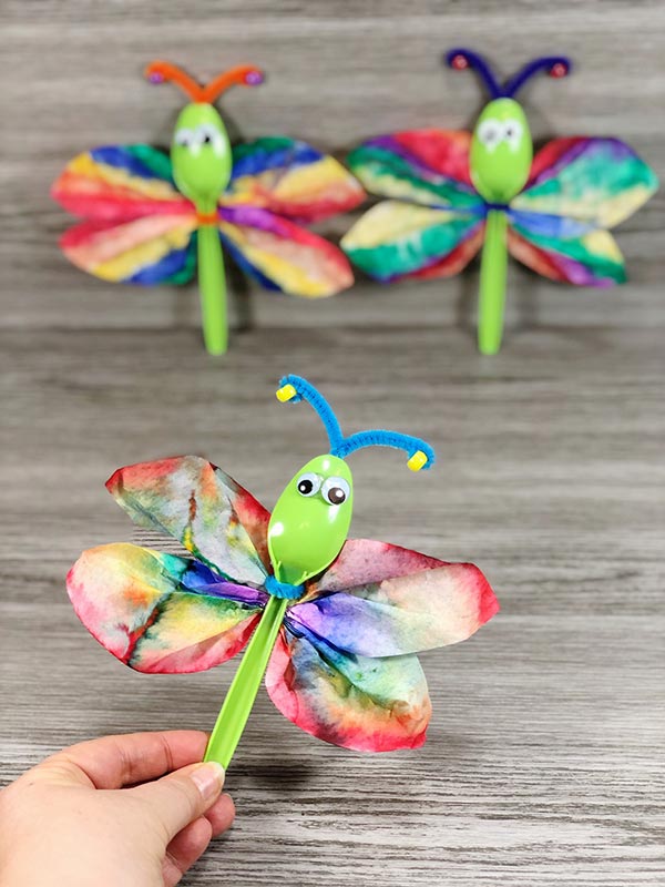 Woman's hand holding up a coffee filter dragonfly and two more in the background on a gray wood backdrop.