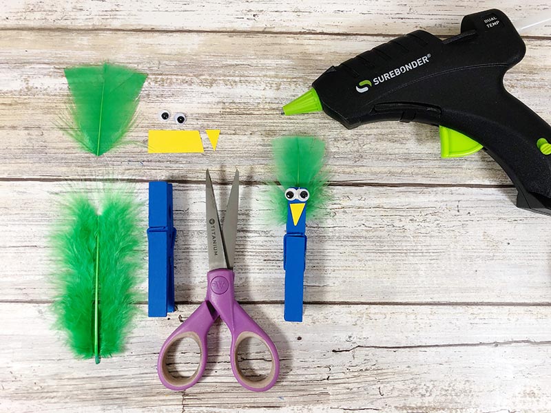 Green feather, blue clothespin, eyes and yellow triangle laying next to assembled peacock clothespin body with glue gun.