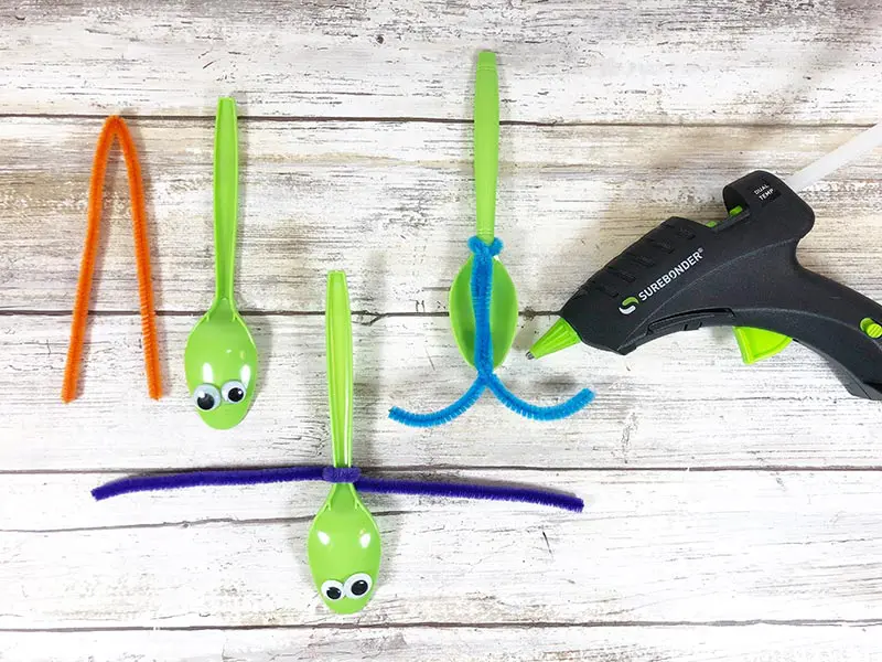 Three green plastic spoons with googly eyes laying next to each other showing steps of how to attach chenille stem to create antenna for dragonfly.