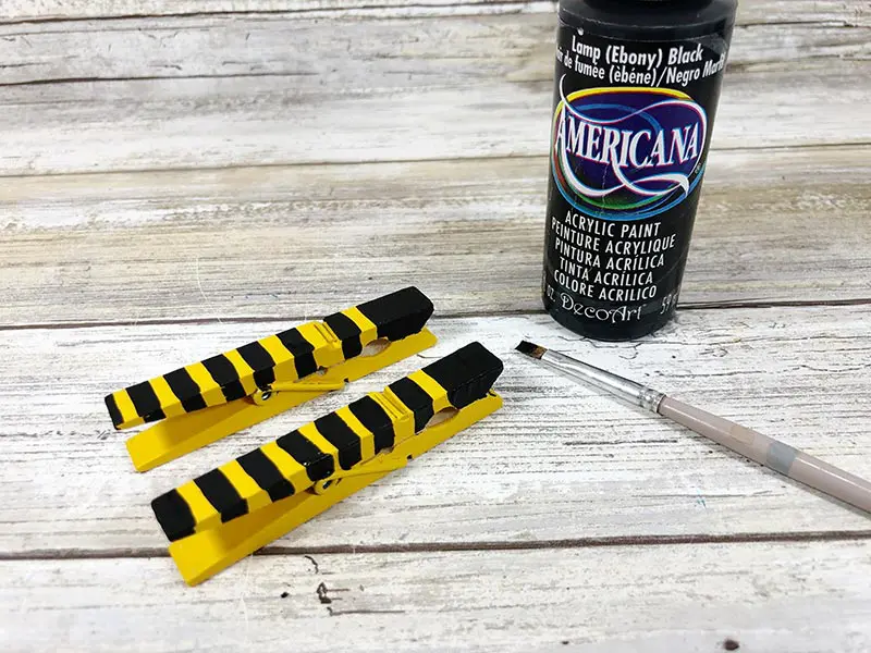 Two clothespins painted yellow with black stripes next to paintbrush and black paint.