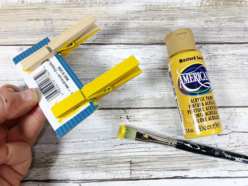 Two wooden clothespins clipped to small piece of cardboard with hand holding the cardboard. One clothespin is painted yellow and the other is unpainted. Paintbrush and yellow paint lay nearby.