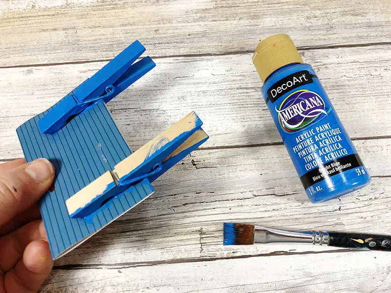 Hand holding piece of cardboard with two wooden clothespins clipped to it. One clothespin is painted blue and the other is in process of being painted. Paint brush and bottle of blue paint laying on white wooden backdrop.