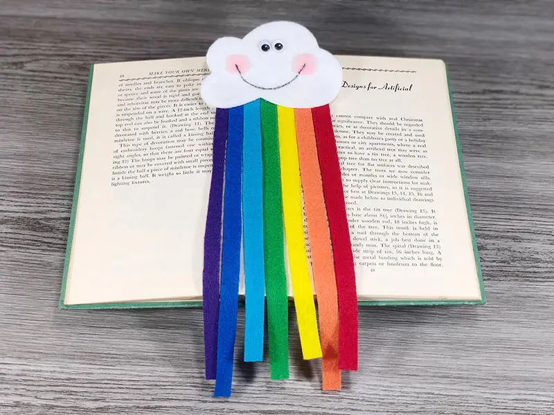 Felt cloud with smiley face and felt rainbow strips coming out of it laying on top of an open book.
