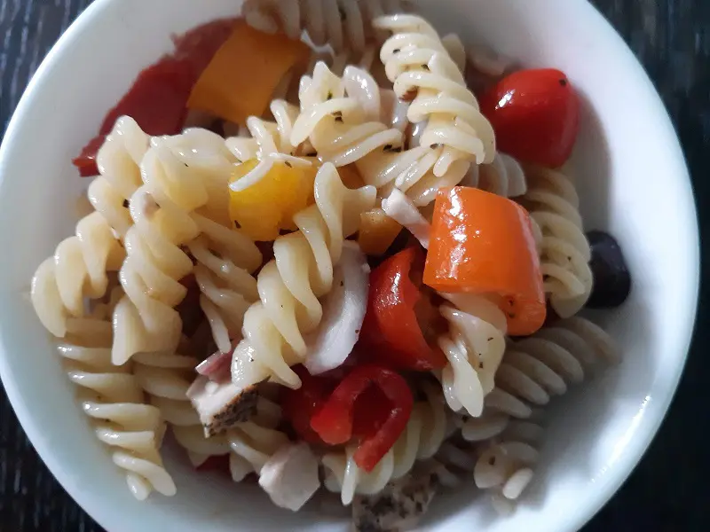 Close up overhead view of white bowl filled with cold pasta salad with noodles, peppers, chicken, pepperoni, and grape tomatoes.
