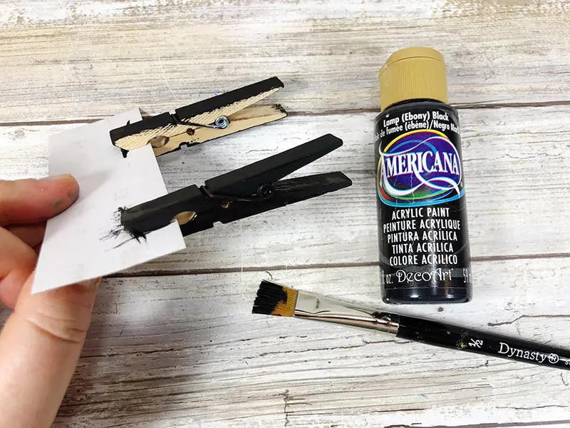 Painting two clothespins black while clipped to a piece of cardboard.