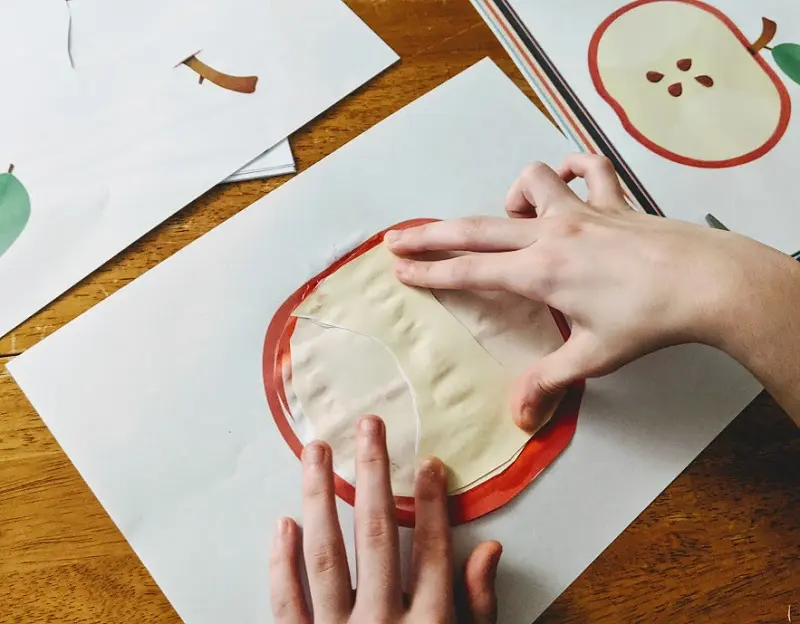 White girl's hands gluing cut out pieces of an apple onto paper.