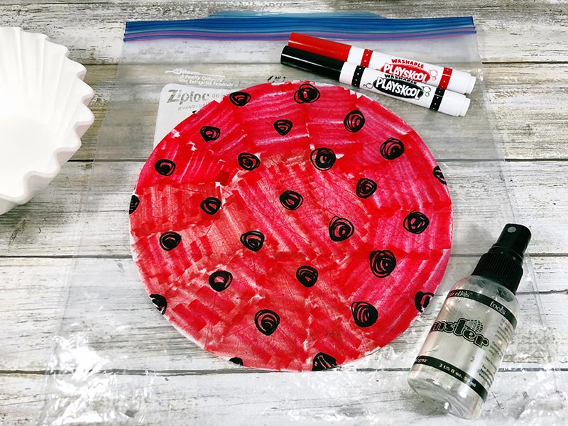 Coffee filter colored with red marker and black circles next to markers, water bottle, and on a plastic bag.