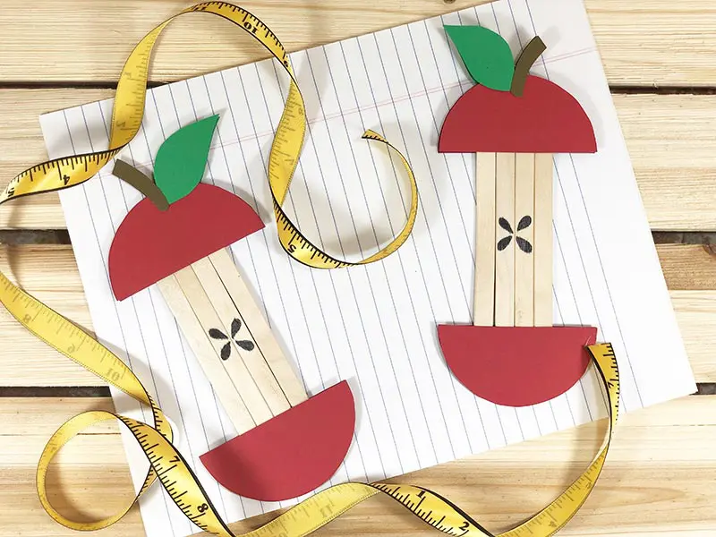 Two popsicle stick apple crafts completely assembled next to soft yellow measuring tape and laying on lined paper.