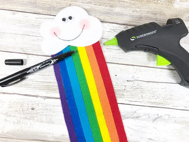 Creating adorable kawaii face on felt cloud with rainbow strips glued to it. Black marker and hot glue gun laying next to project.