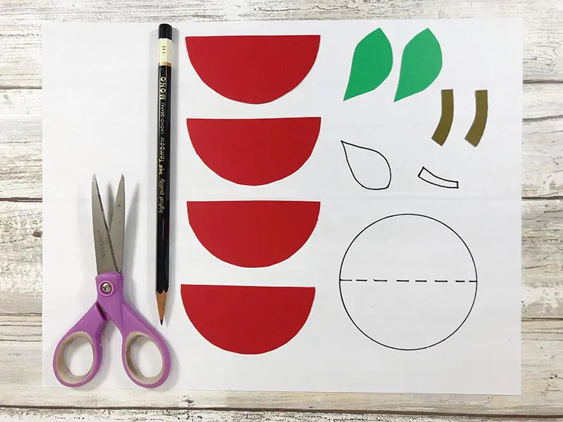 Purple handled scissors and pencil laying next to apple pieces cut out of cardstock paper next to craft pattern.