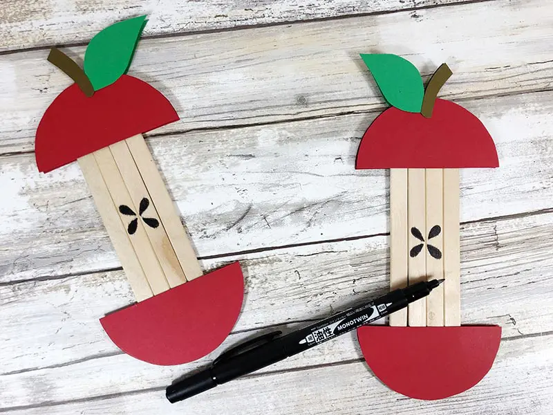 Two completely assembled craft stick apples showing black marker used to draw on four teardrop shaped seeds for core.
