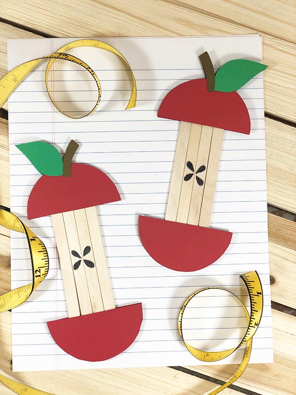 Two red apple cores made with paper and popsicle sticks laying on loose leaf paper with a soft measuring tape curled around the edges.