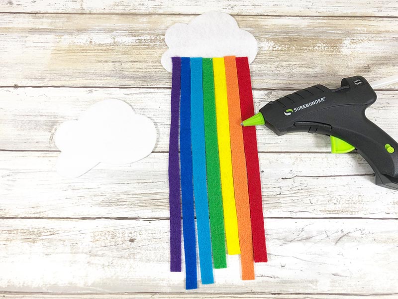 Felt strips arranged to make a rainbow glue to back of white felt cloud laying next to hot glue gun.