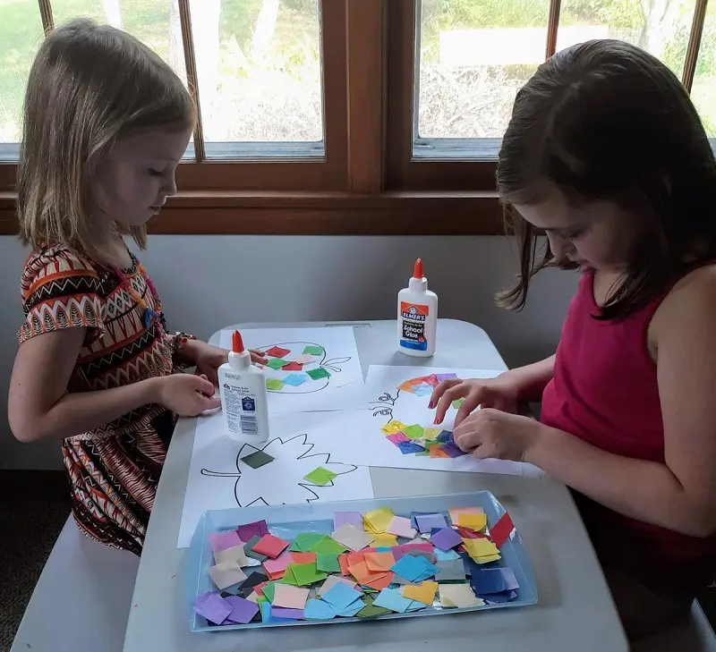 Two young white girls sitting across from each other at a table gluing tissue paper squares to the fall craft templates.