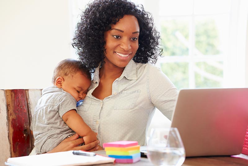 Smiling Black mom holding sleeping baby while working on laptop.