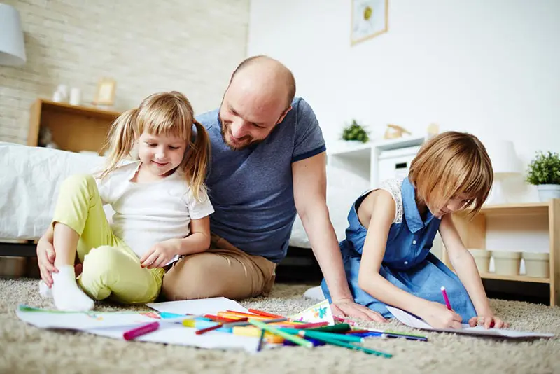White dad sitting on floor with two daughters smiling and drawing.